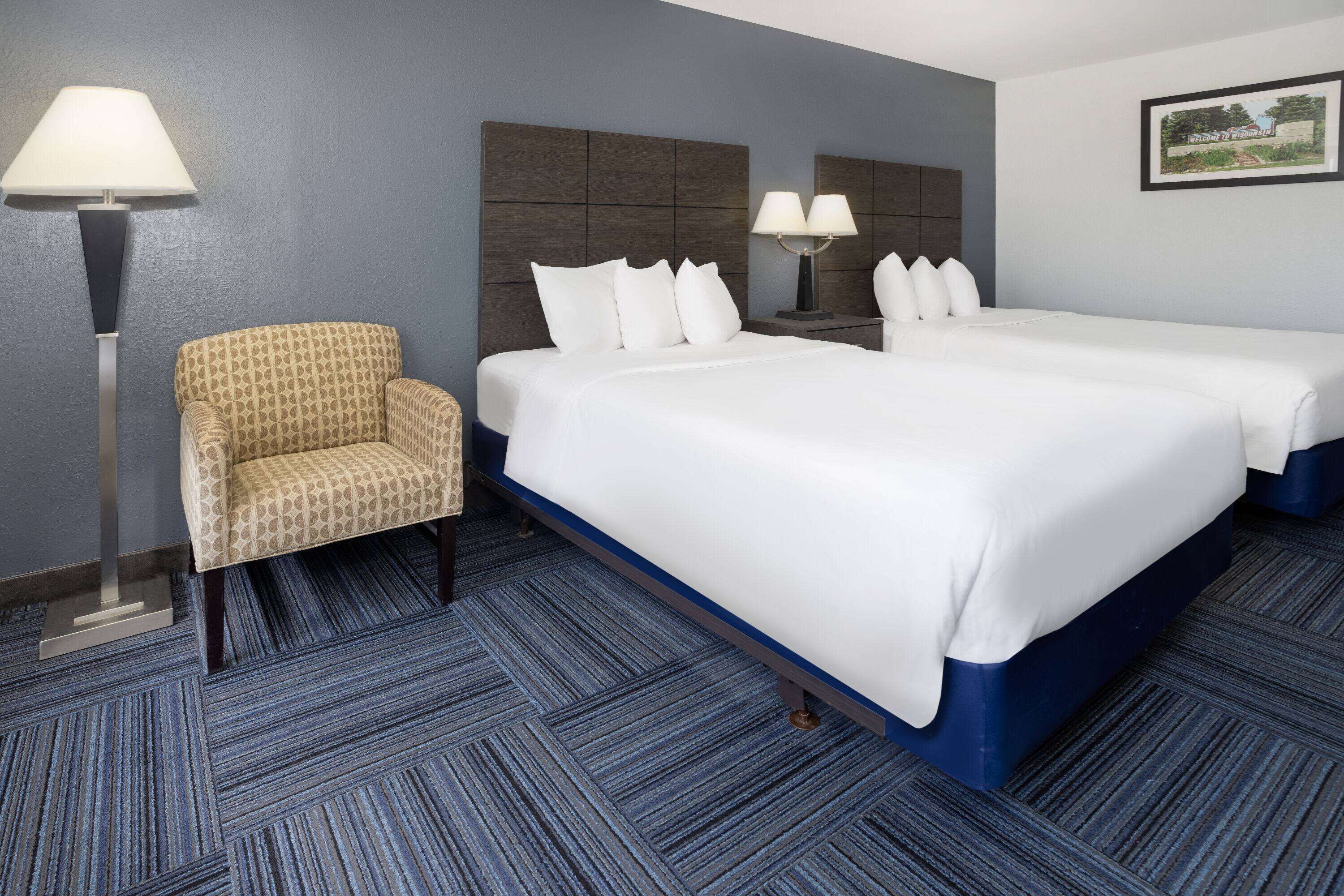A hotel room with two beds, a patterned armchair, a floor lamp, and a framed welcome sign.