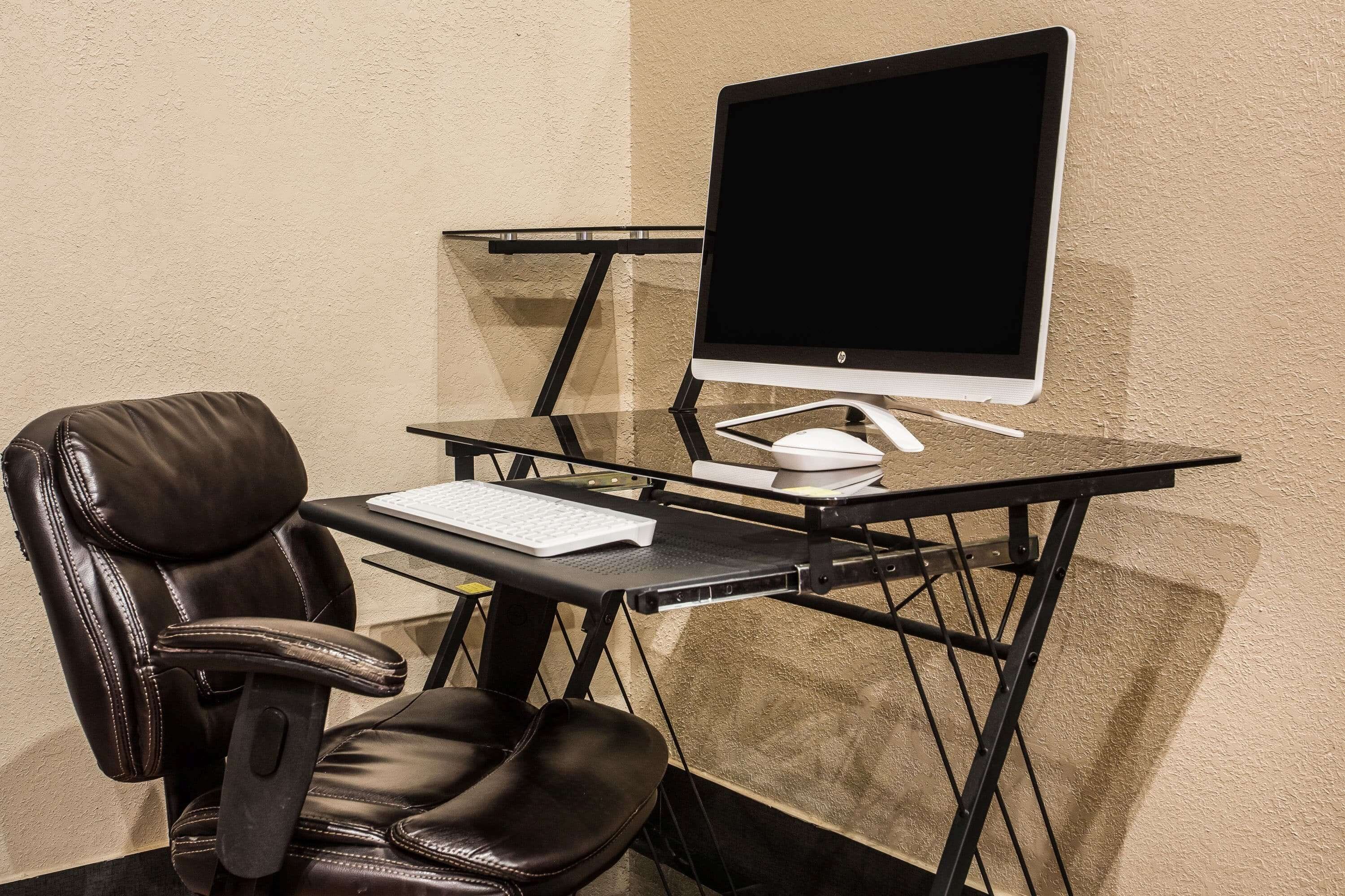 A modern office chair with a high backrest and armrests, a glass desk with a monitor, keyboard, and mouse, and a carpeted floor.