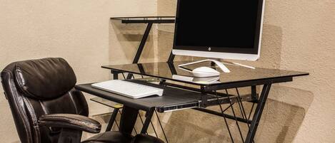 A modern office chair with a high backrest and armrests, a glass desk with a monitor, keyboard, and mouse, and a carpeted floor.
