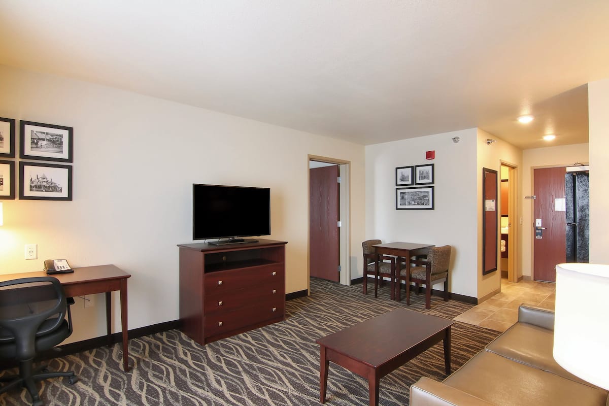 A hotel room with a flat-screen TV, a desk with a telephone, a chair, a dining table with chairs, and a carpeted floor.