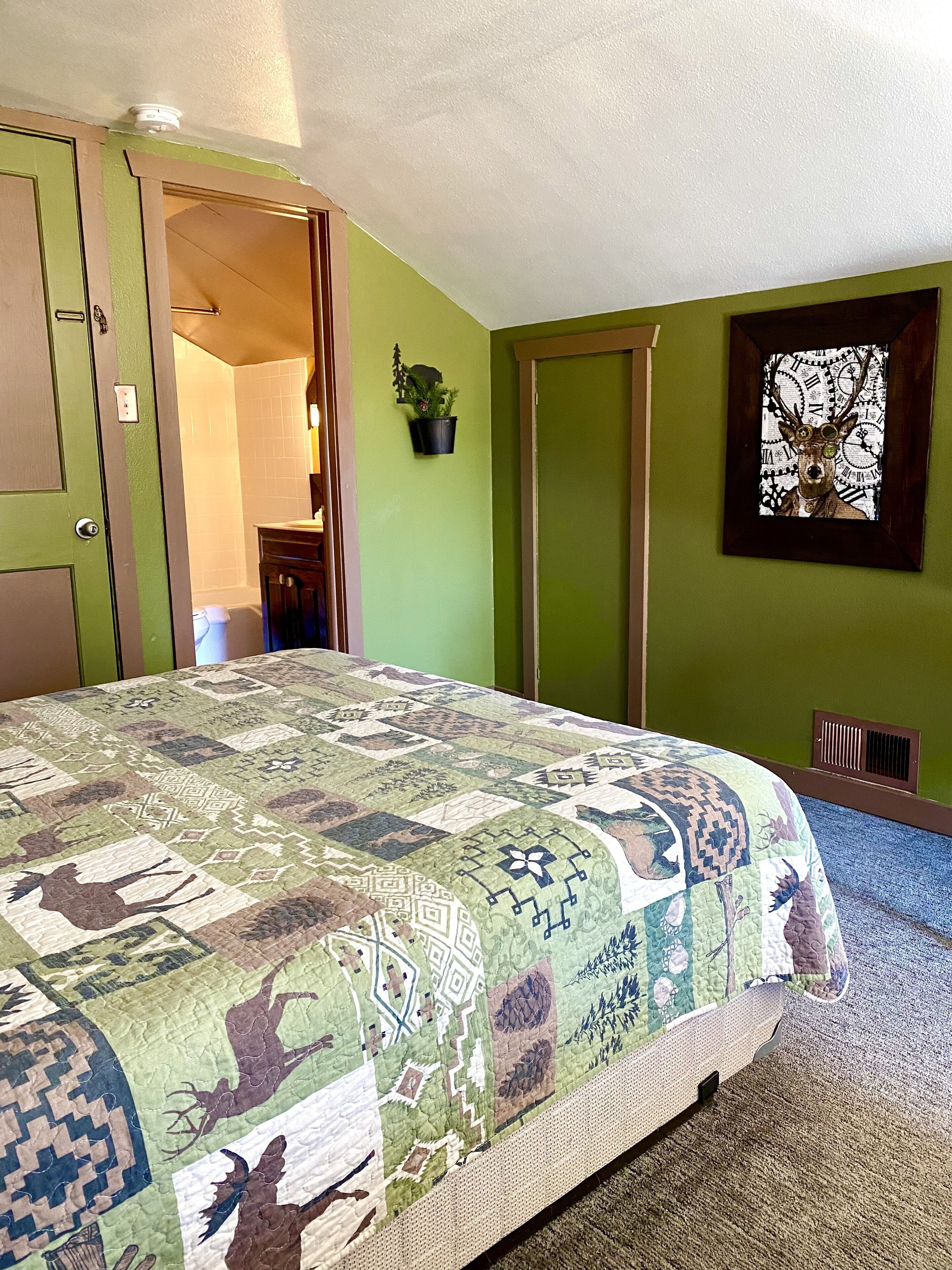 A bedroom with a large bed, a green wall, a window with a deer picture, and a door leading to another room.