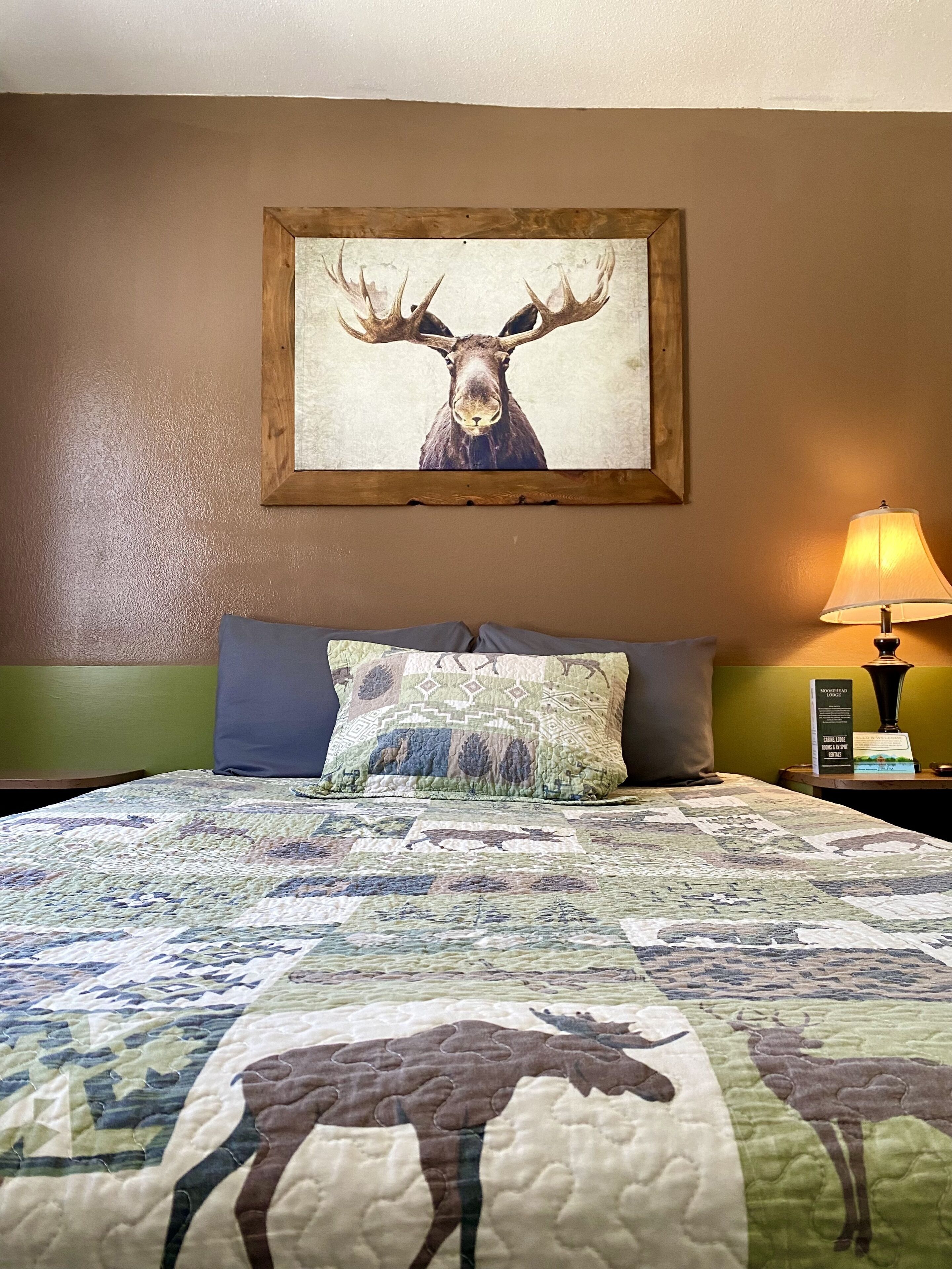 A bedroom with a large bed, a moose painting on the wall, and a lamp on the nightstand.