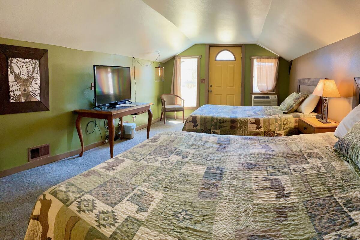 Un dormitorio con una cama grande, un televisor sobre un mueble de madera, una silla y una puerta que da a otra habitación.