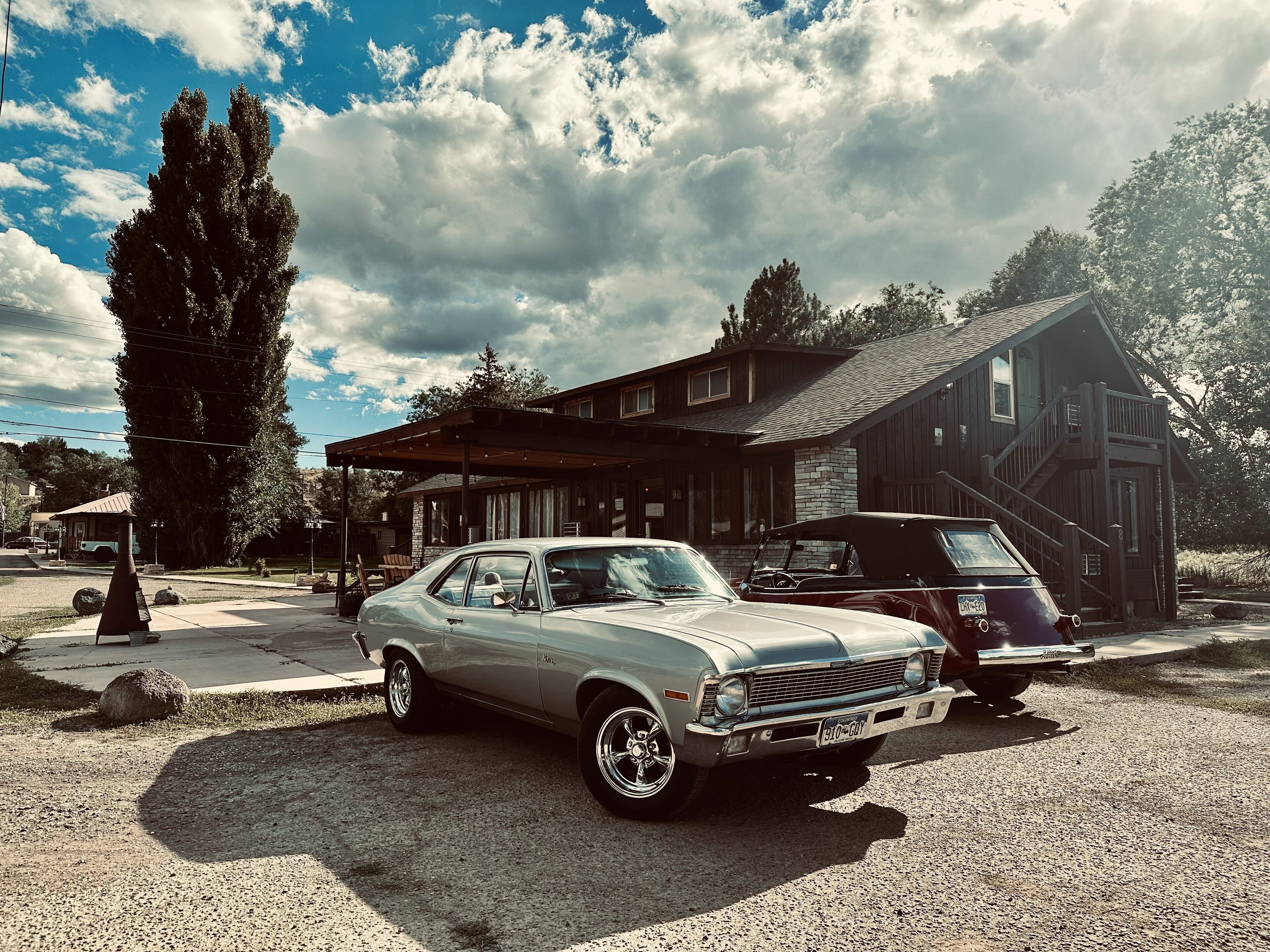 Une voiture ancienne est garée devant une maison en bois dotée d’un porche et d’un escalier.
