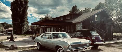 Une voiture ancienne est garée devant une maison en bois dotée d’un porche et d’un escalier.