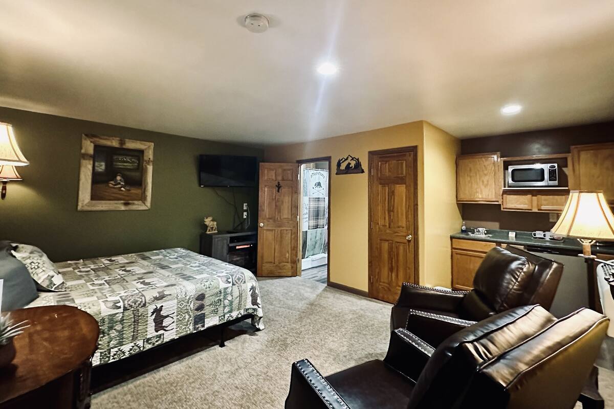 Un dormitorio con cama, una silla, televisor, puerta de madera y una zona de cocina con microondas.