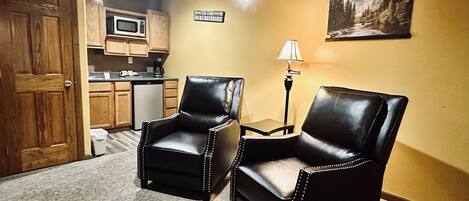 A room with two leather chairs, a kitchen area, a door, a lamp, and a framed picture on the wall.