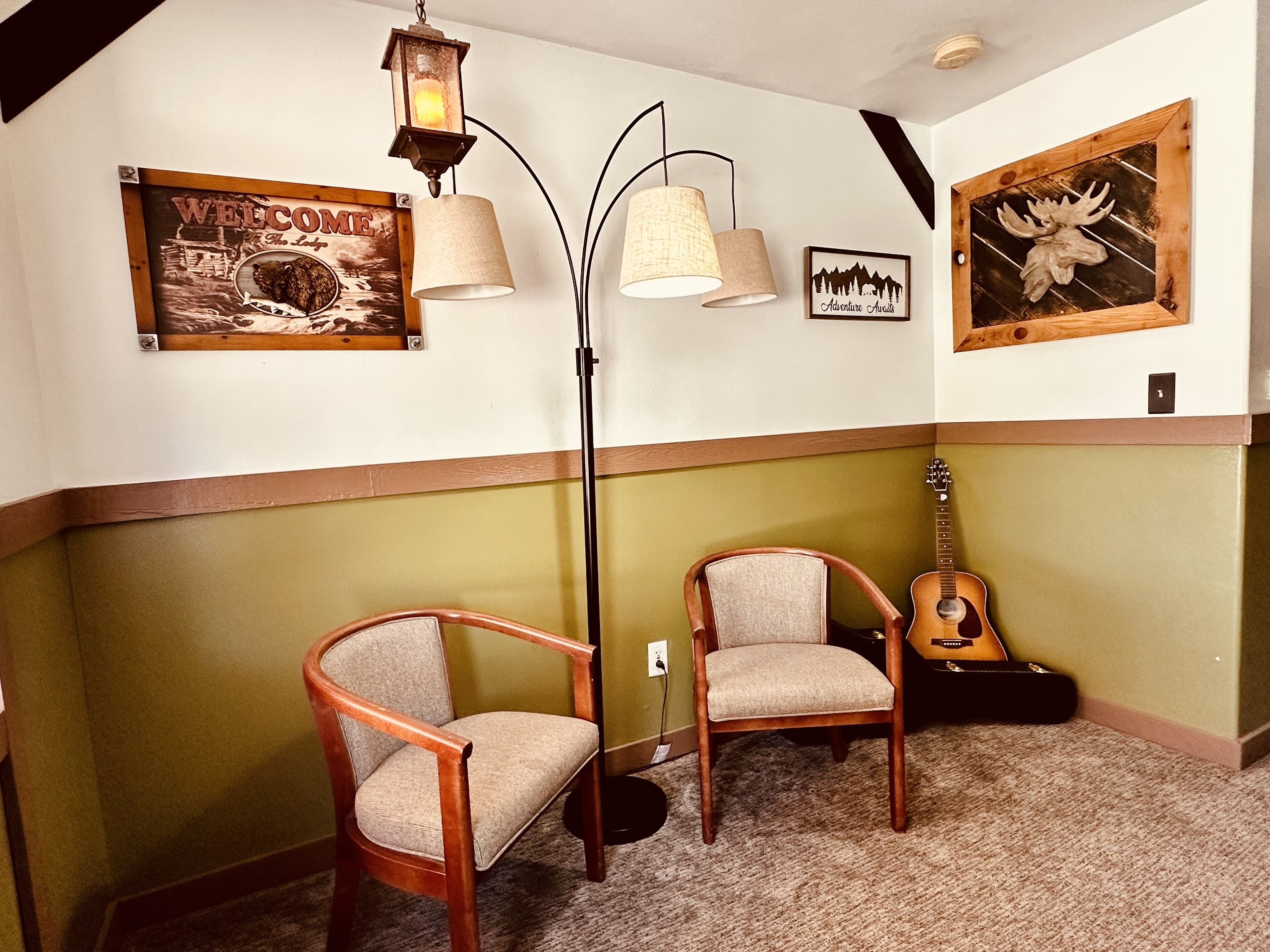 A room with two wooden chairs, a guitar, a floor lamp, and wall art including a 'Welcome' sign and a moose head.