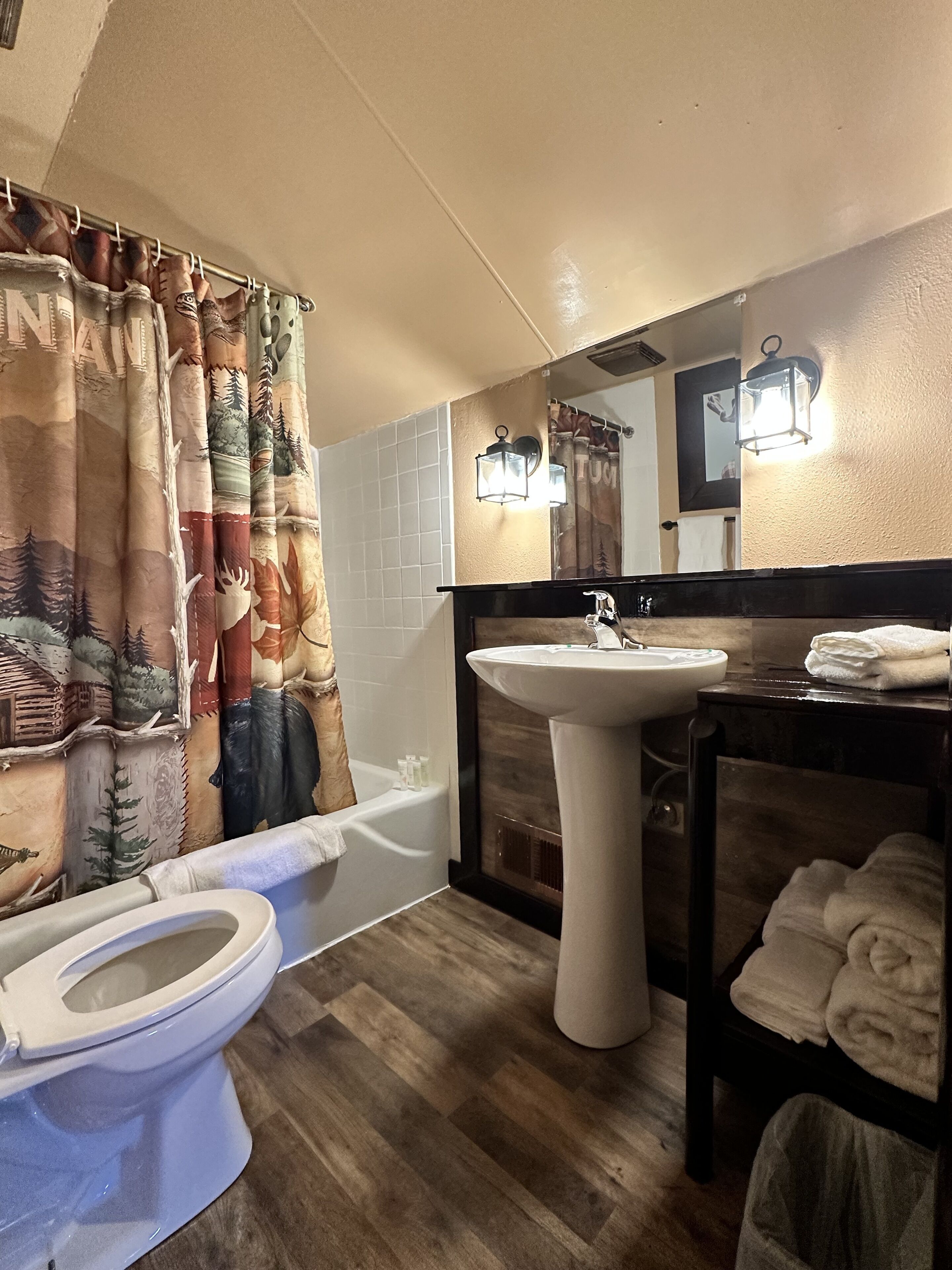 A bathroom with a wooden floor, a white sink, a toilet, a bathtub, and a shower curtain with a forest design.