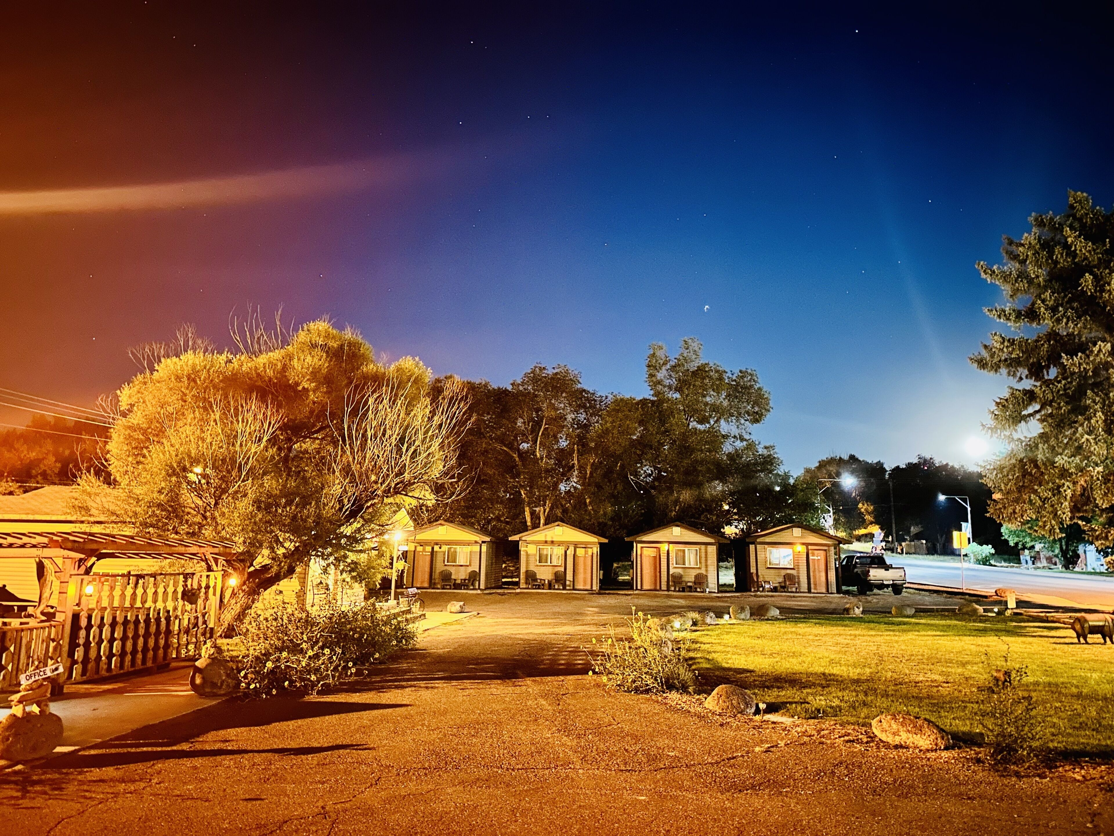 A row of single-story cabins with a paved driveway leading to the entrance, surrounded by trees and a well-lit street.