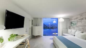 Ein modernes Hotelzimmer mit einem großen Bett, einem Schreibtisch mit Flachbildfernseher, einem kleinen Tisch und Blick auf die Stadt durch eine Glasdoor.