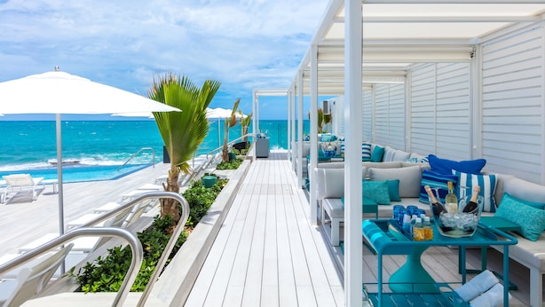 A deck with white railings, a white umbrella, and a seating area with blue cushions and a table with drinks.