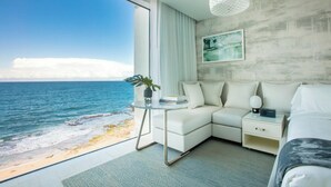 A modern hotel room with a large window overlooking the sea, a white sofa, a small table with a plant, and a framed picture on the wall.