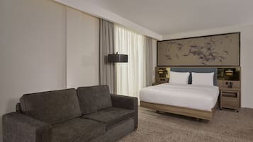 A hotel room with a large bed, a sofa, a nightstand with a lamp, and a wall-mounted artwork.