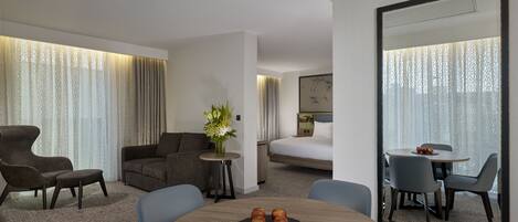 Una habitación de hotel moderna con sofá, una mesa de comedor con manzanas, una pequeña mesita con un jarrón y un espejo grande.