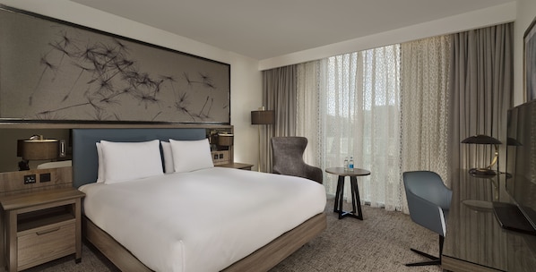 Une chambre d’hôtel avec un grand lit, un bureau, une chaise, une petite table et une grande fenêtre avec des rideaux.