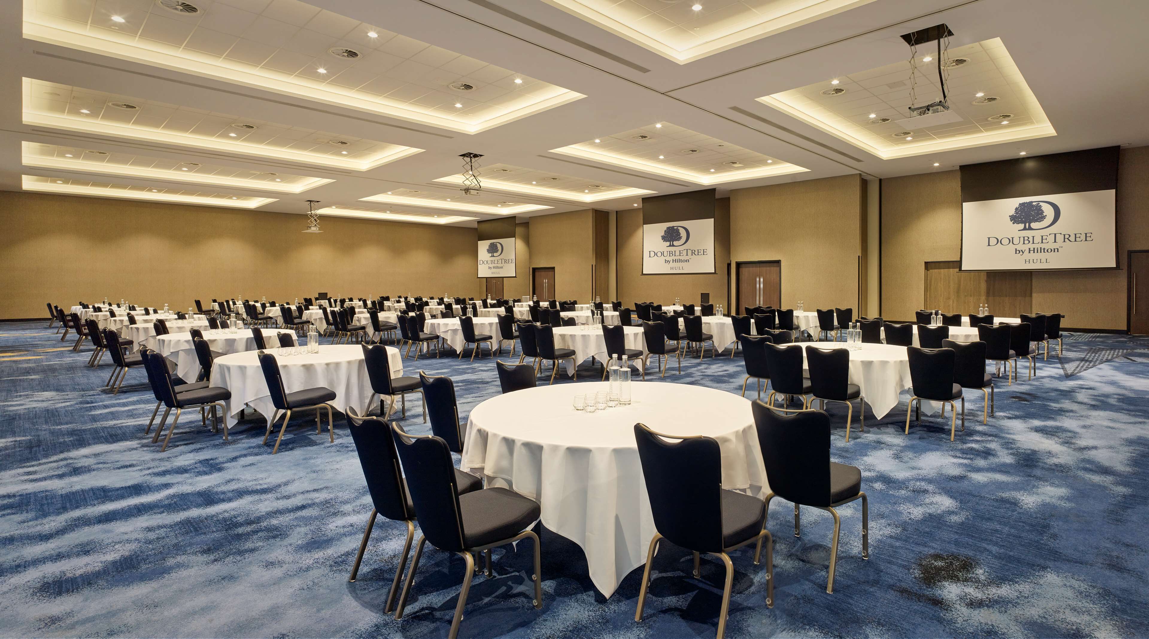 Une grande salle de conférence avec des tables et des chaises rondes, une moquette bleue et des luminaires au plafond.
