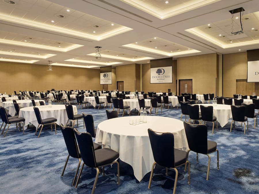 Un amplio salón de conferencias con mesas y sillas redondas, alfombra azul e iluminación en el techo.