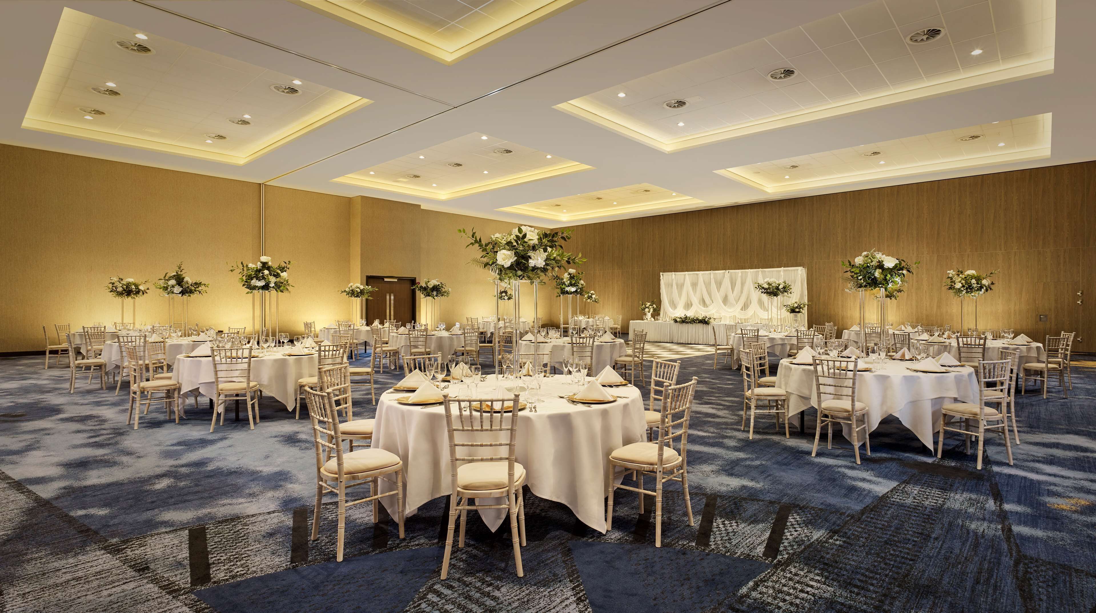 Un amplio salón de banquetes con mesas redondas, manteles blancos y altos arreglos florales.