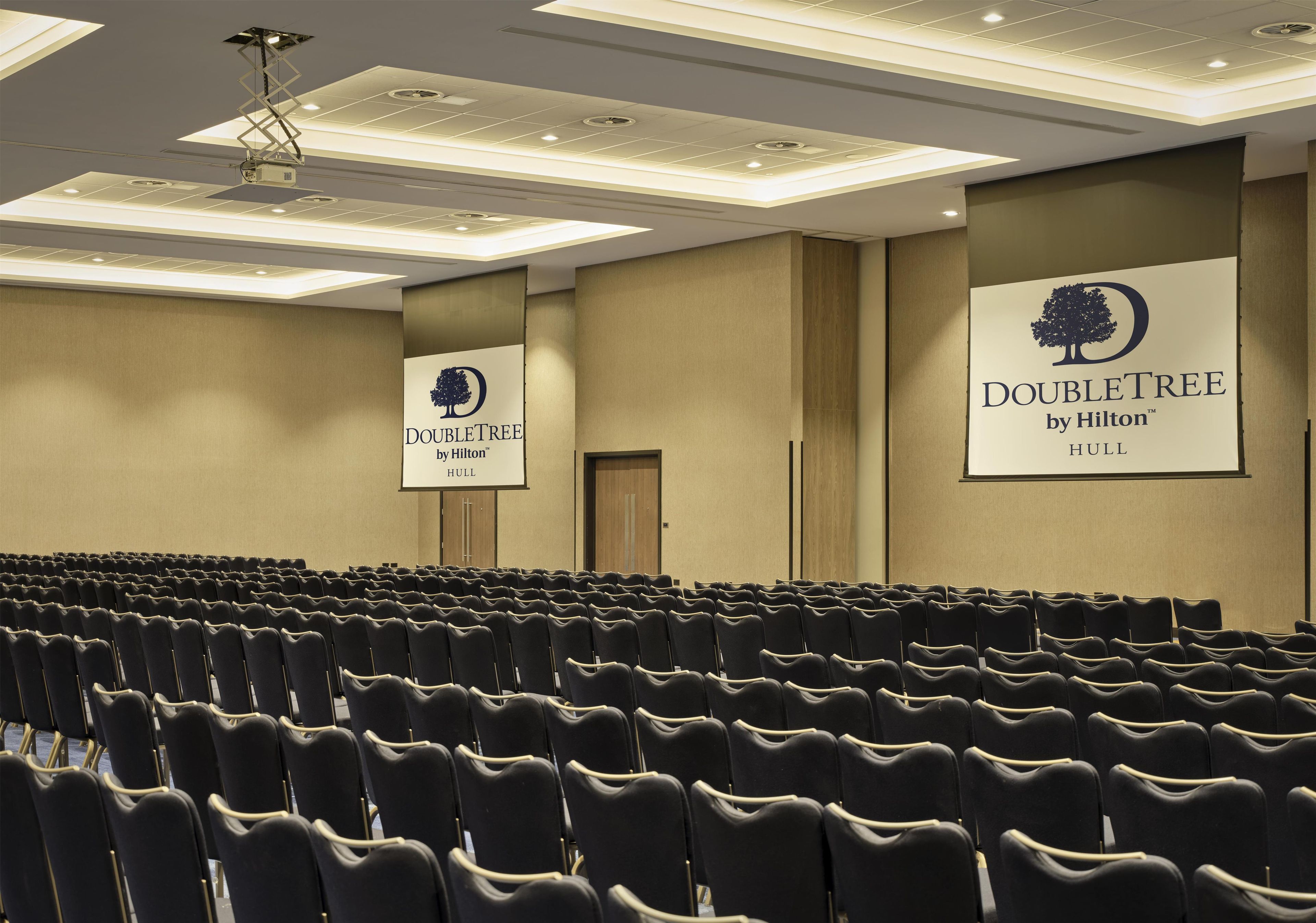 Ein Konferenzraum mit Reihen schwarzer Stühle, einer Decke mit eingelassener Beleuchtung und zwei großen Bannern für DoubleTree by Hilton Hull.