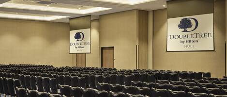 Ein Konferenzraum mit Reihen schwarzer Stühle, einer Decke mit eingelassener Beleuchtung und zwei großen Bannern für DoubleTree by Hilton Hull.