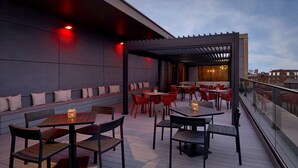 An outdoor patio with wooden tables and chairs, a pergola, and a view of the city skyline.