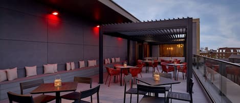 Un patio al aire libre con mesas y sillas de madera, una pérgola y vistas del horizonte de la ciudad.