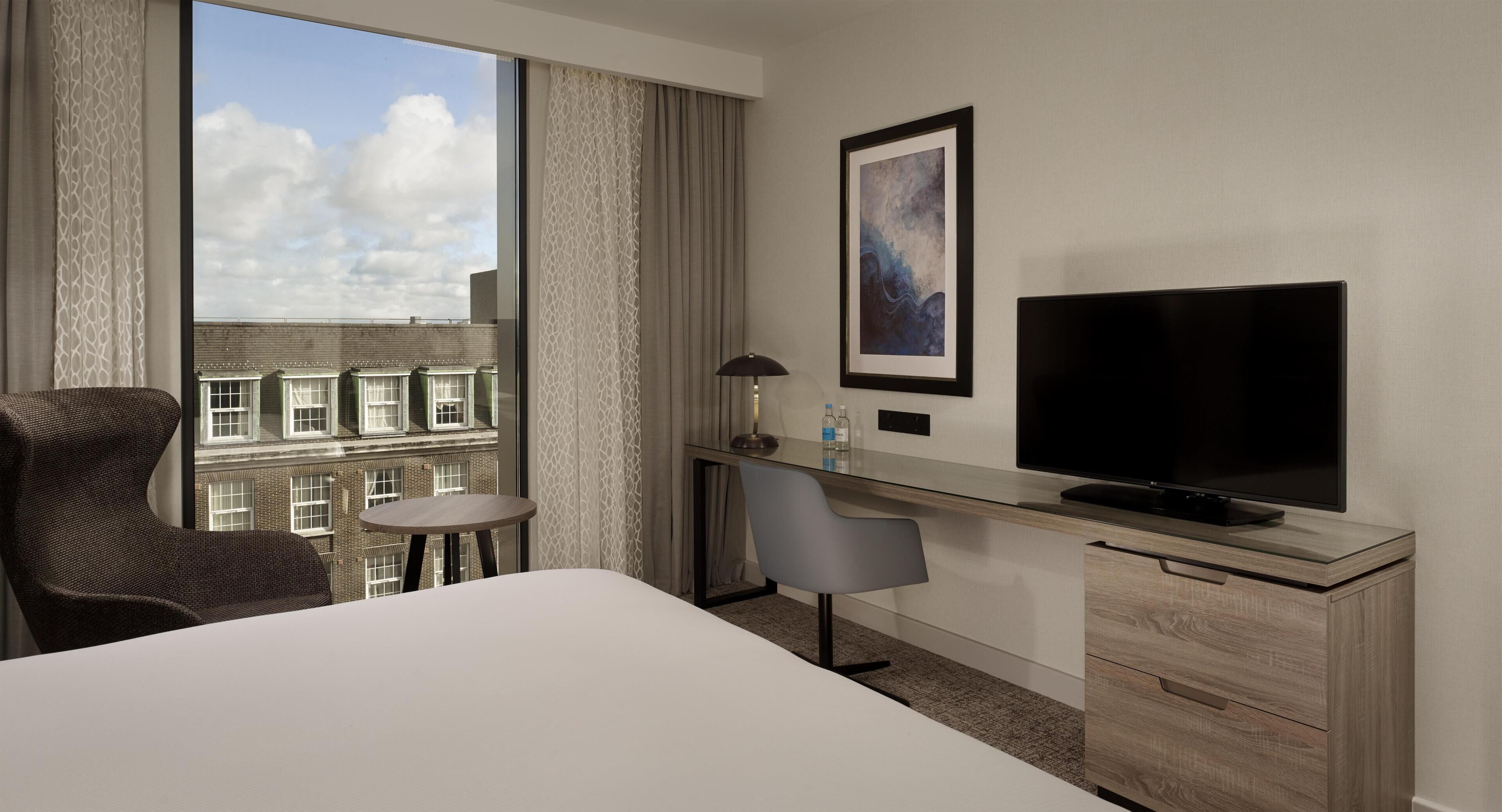 A hotel room with a bed, a desk with a TV, a chair, a small table, and a window with a view of buildings.