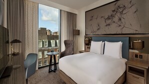 A hotel room with a large bed, a chair, a small table, and a view of the city through the window.