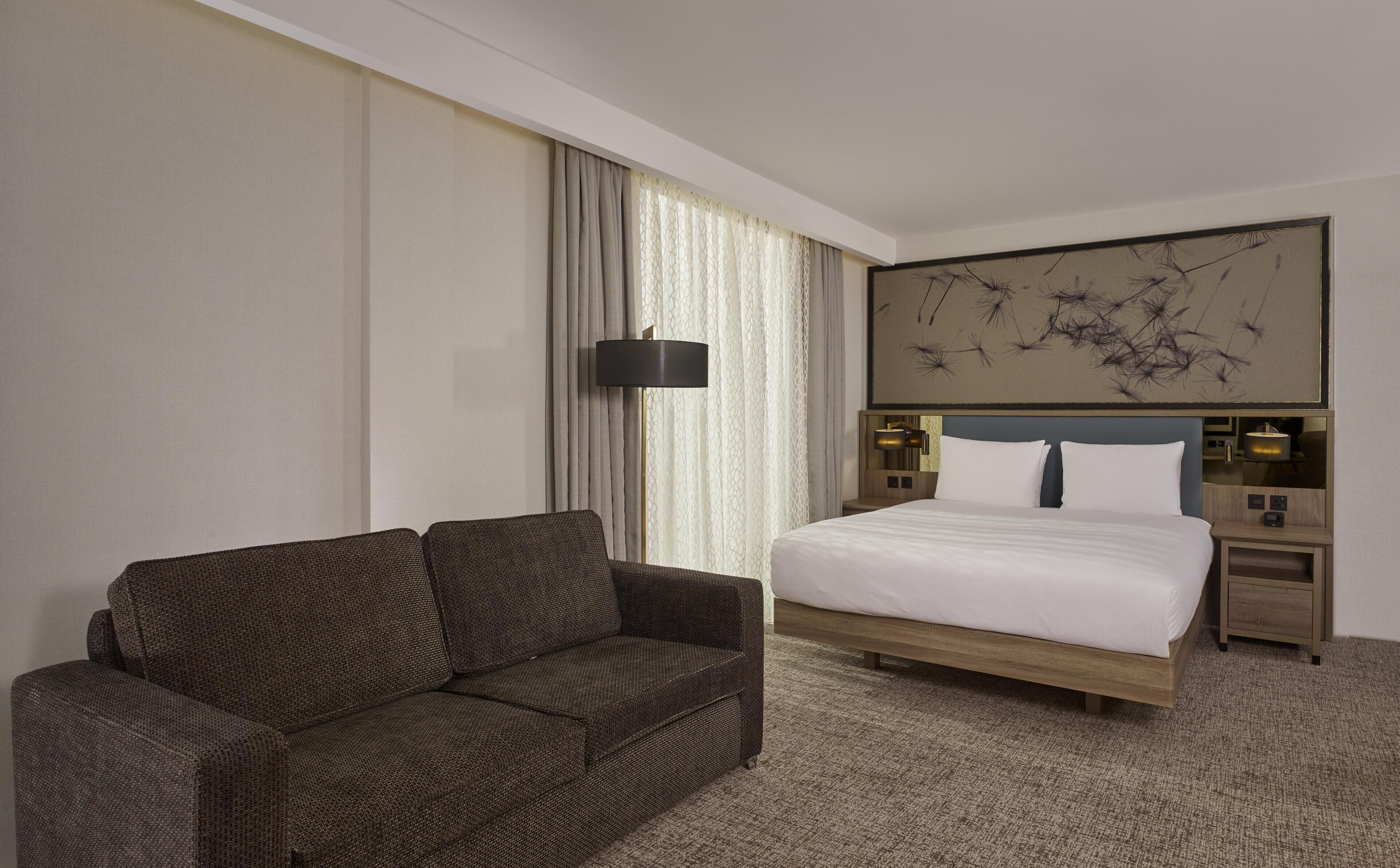 A hotel room with a bed, a sofa, a nightstand, a lamp, and a wall art piece.