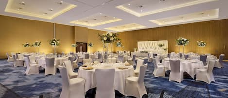 Un amplio salón de banquetes con mesas redondas, sillas blancas y centros de mesa florales.