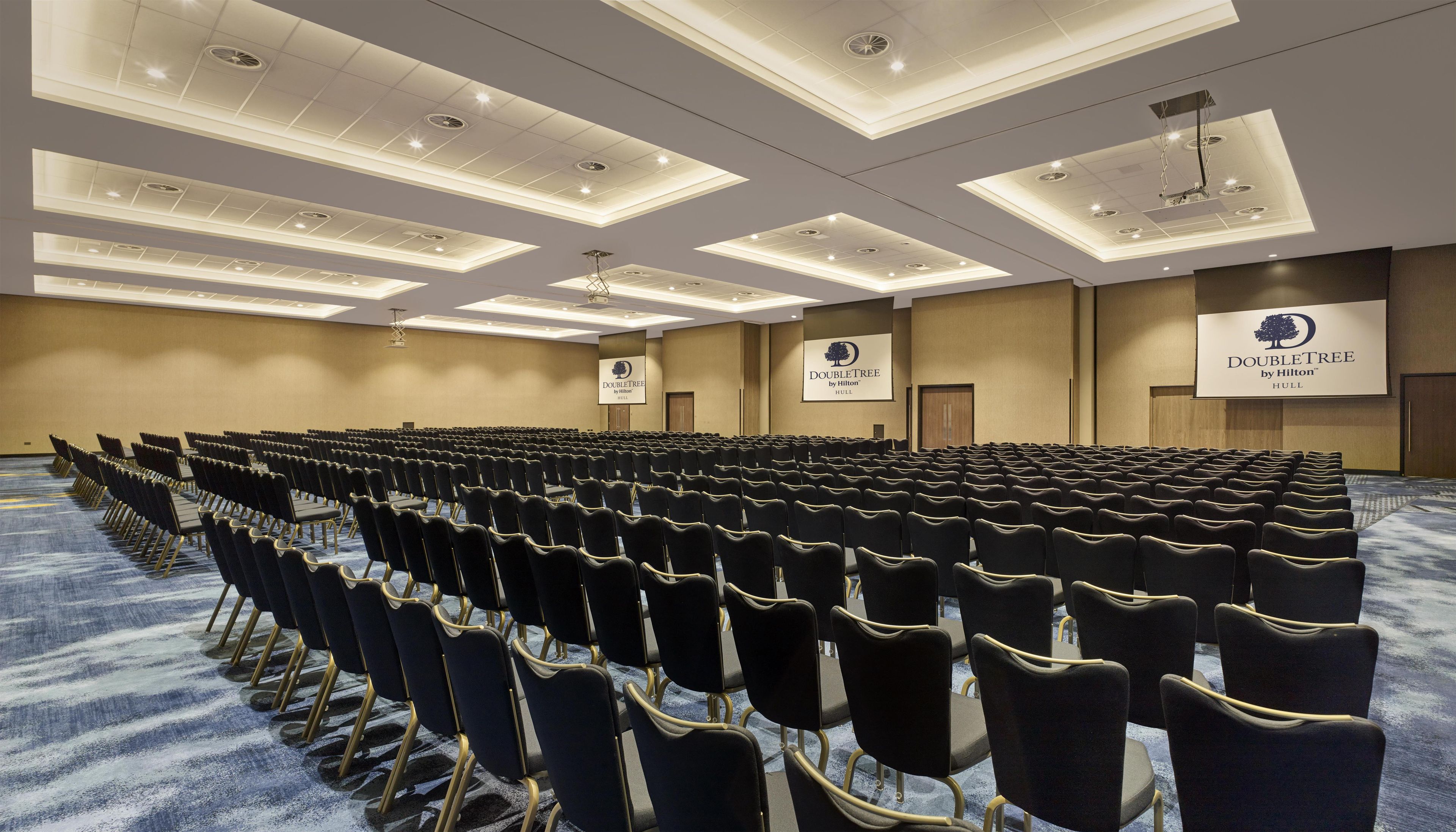 Un salón de conferencias con filas de sillas y una pantalla grande que muestra el logotipo de DoubleTree by Hilton.