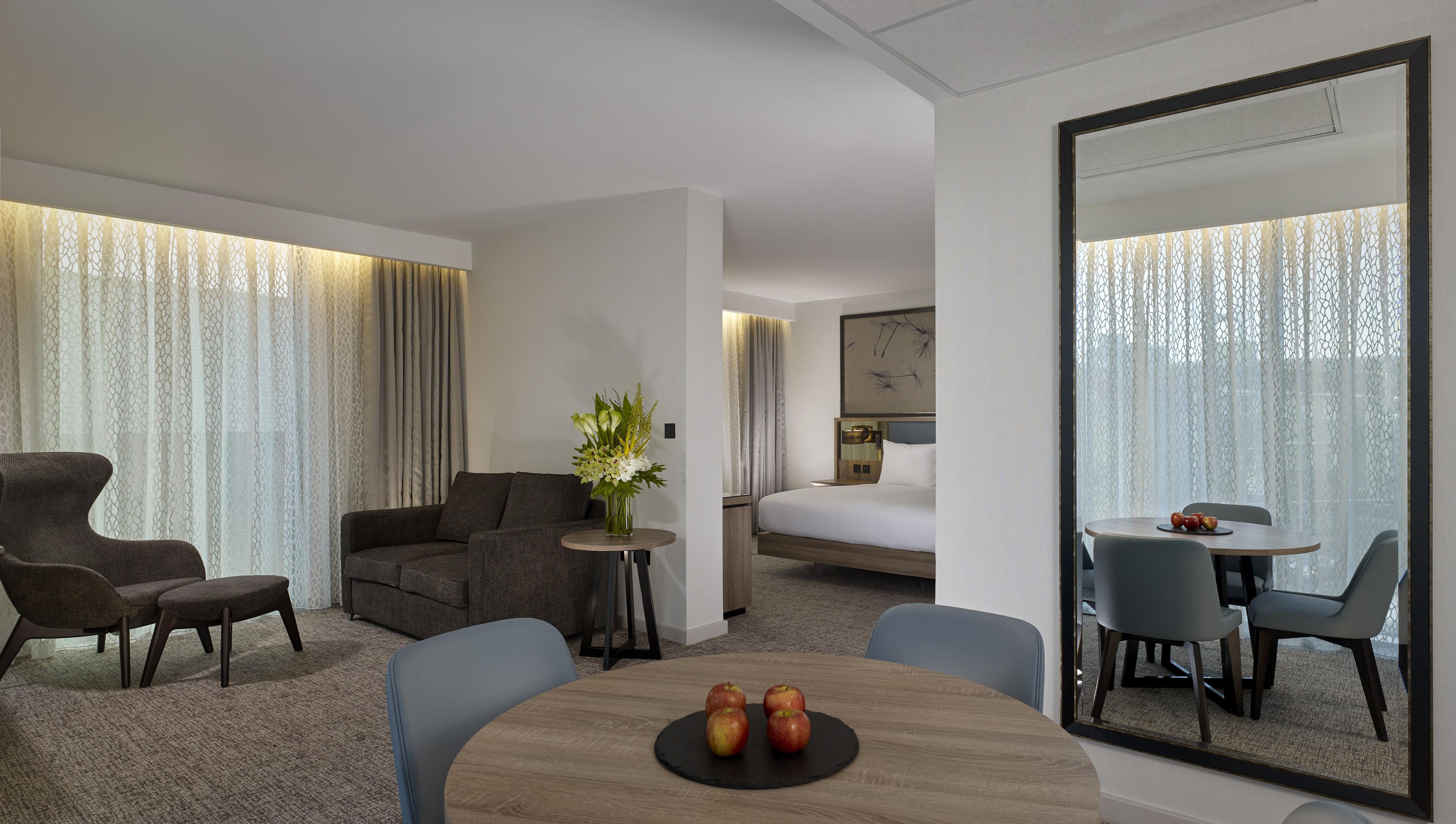 A hotel room with a sofa, a small round table with apples, a dining area with chairs, and a large mirror.