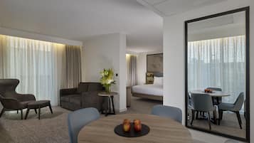 A hotel room with a sofa, a small round table with apples, a dining area with chairs, and a large mirror.
