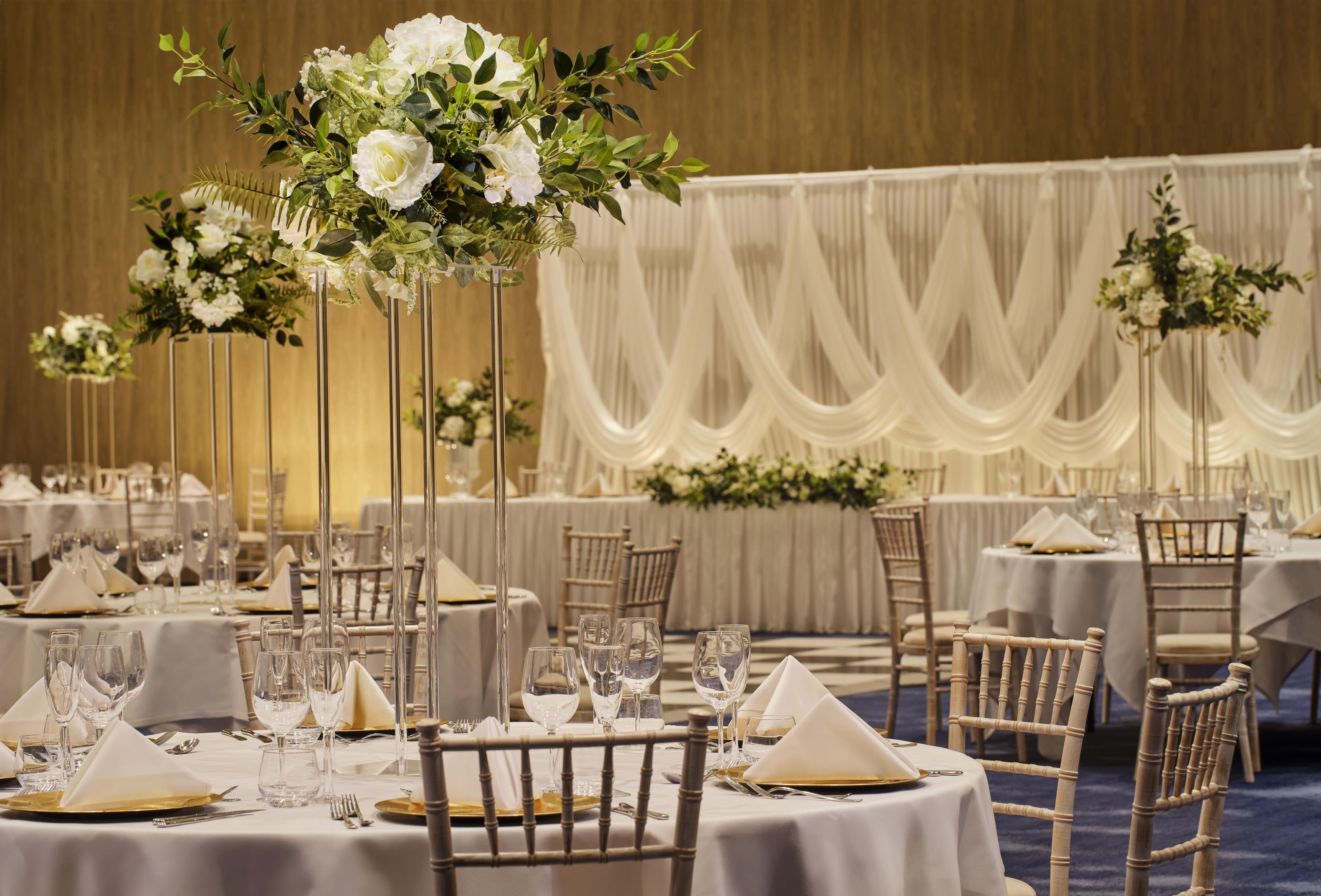 Un salón de banquetes con mesas redondas dispuestas para el servicio de comidas, cada una con mantel blanco, platos base dorados y copas de cristal.