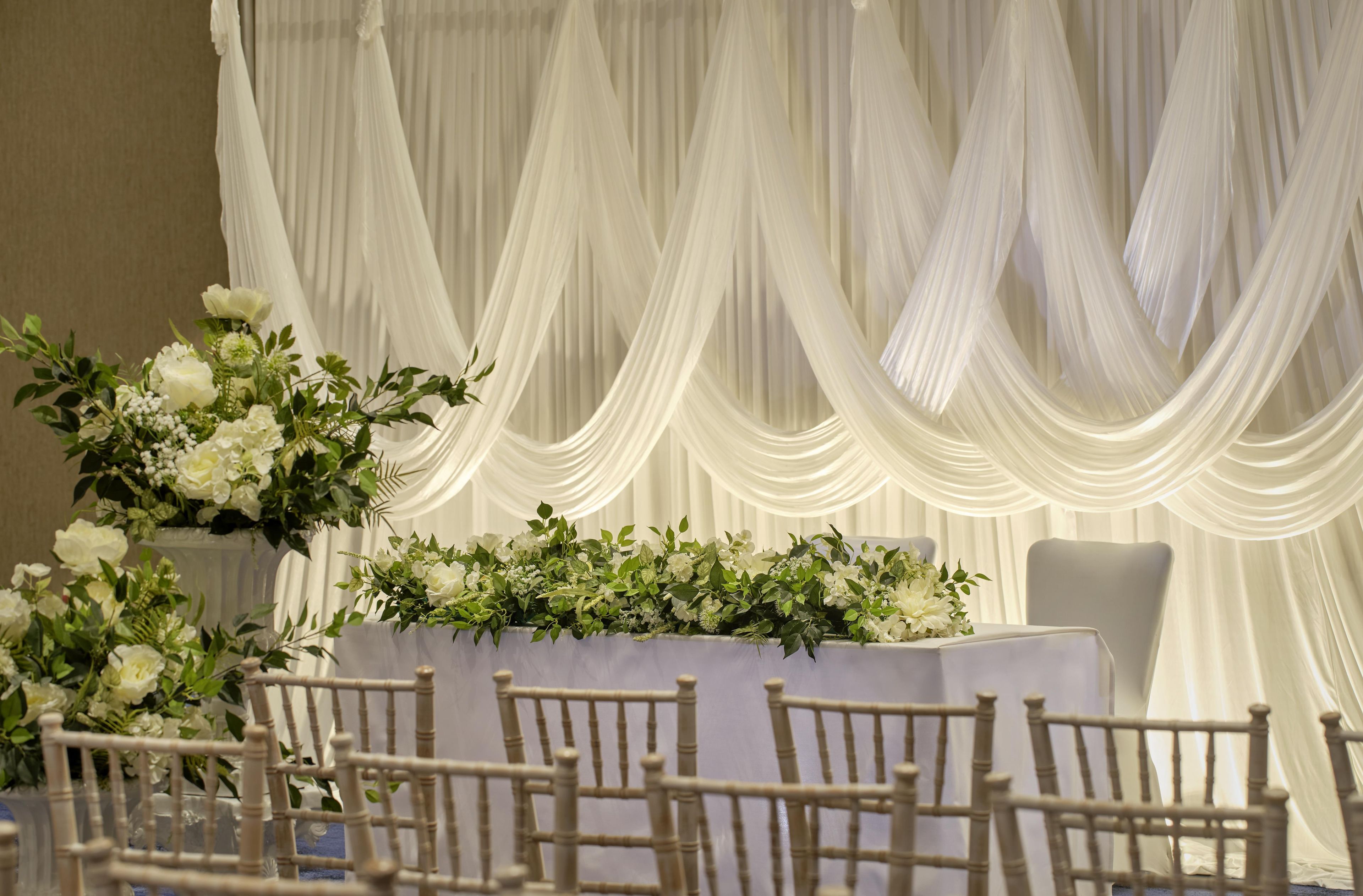 A table set with white tablecloth and chairs, a floral arrangement, and a backdrop of white drapes.