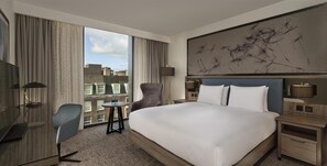 A hotel room with a large bed, a desk, a chair, a small table, a nightstand, a lamp, and a view of buildings through a window.