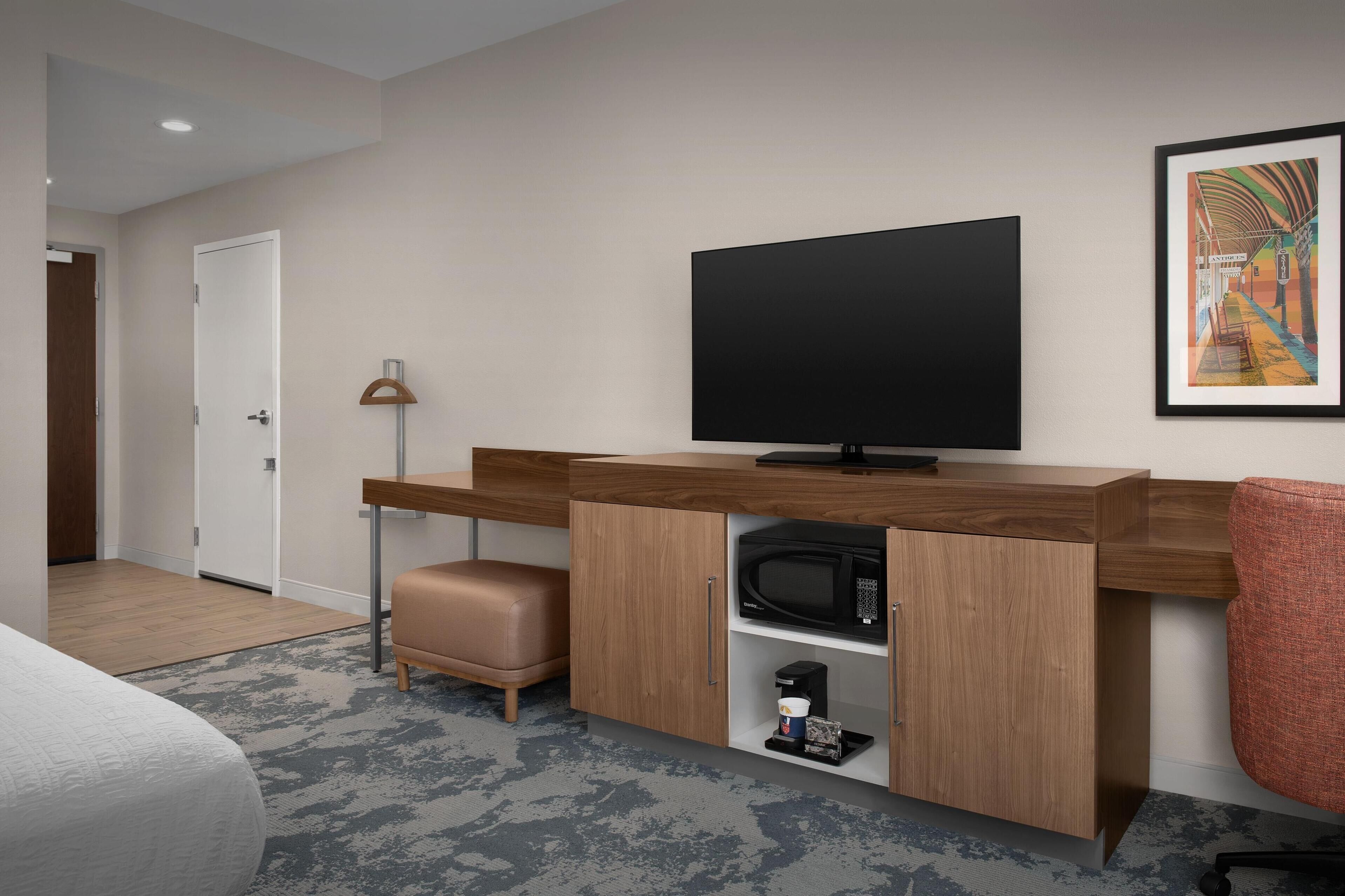 Una moderna camera d'albergo con una scrivania in legno, una TV a schermo piatto, un forno a microonde e un'opera d'arte incorniciata appesa al muro.
