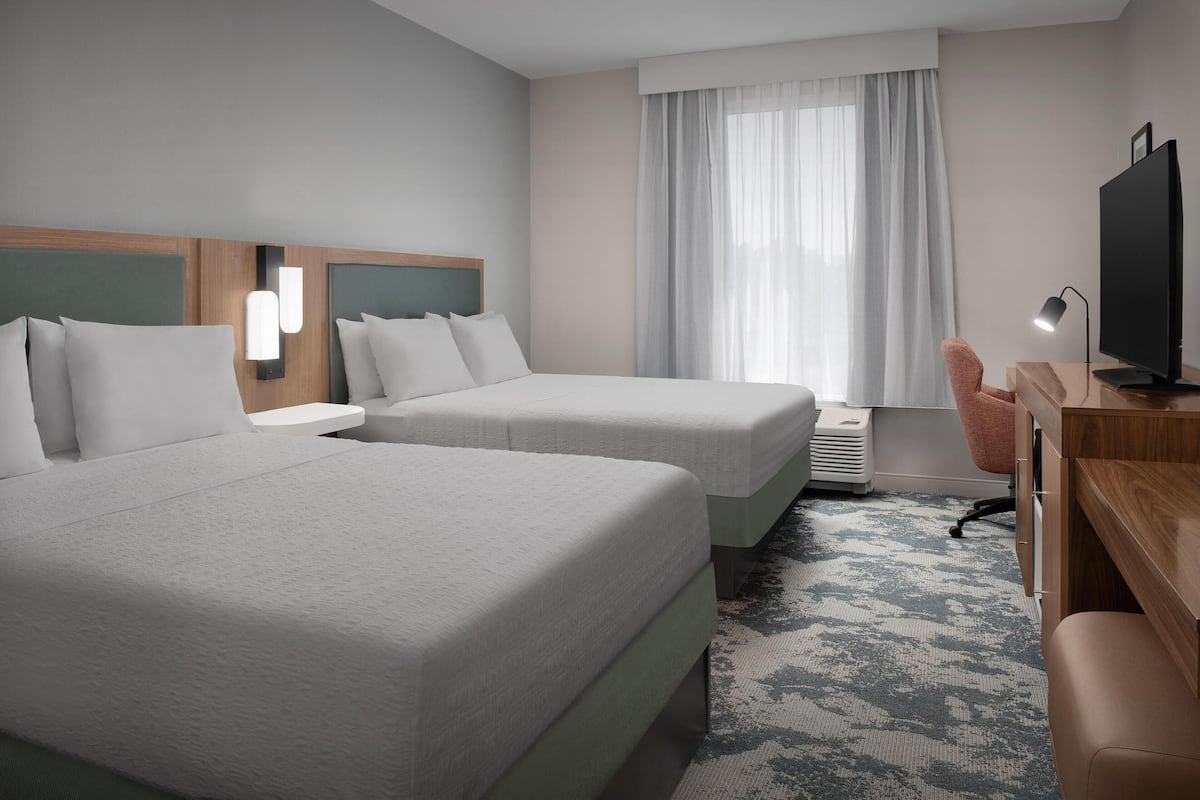 Une chambre d’hôtel avec deux lits, un bureau, une chaise, une télévision et une fenêtre avec des rideaux.