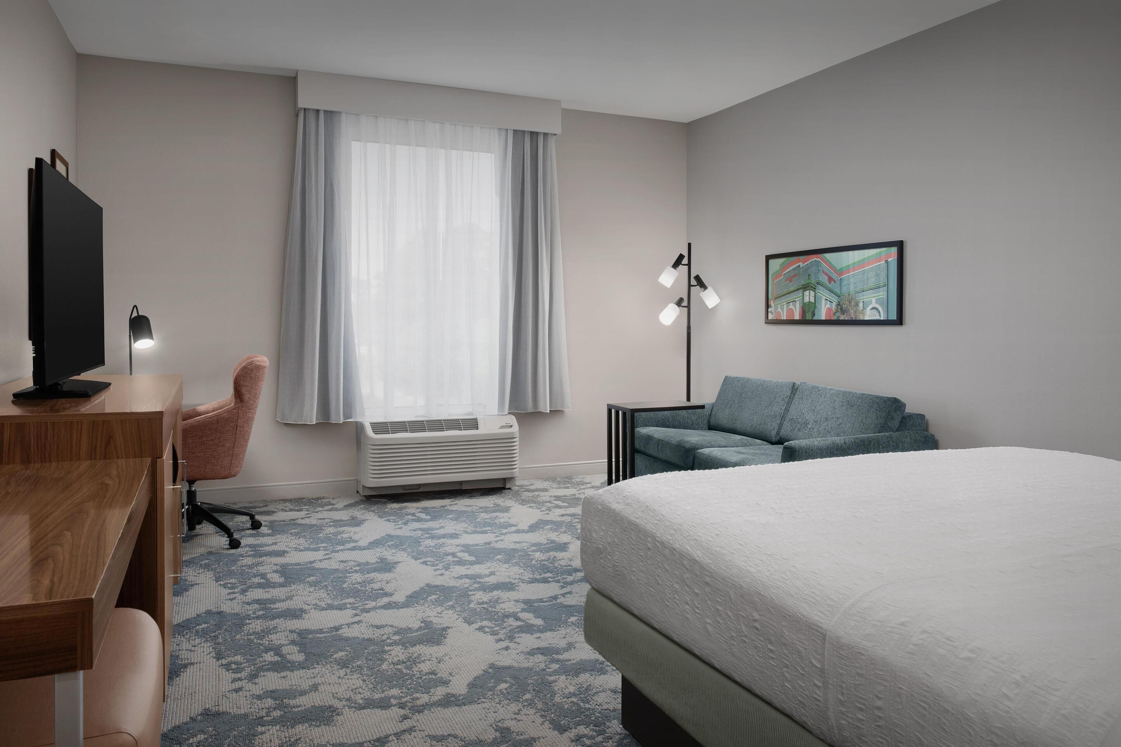 A hotel room with a bed, a desk with a computer, a chair, a standing lamp, and a large window with curtains.