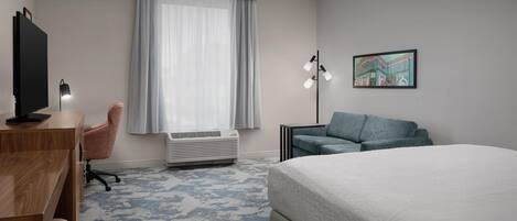 A hotel room with a bed, a desk with a computer, a chair, a standing lamp, and a large window with curtains.