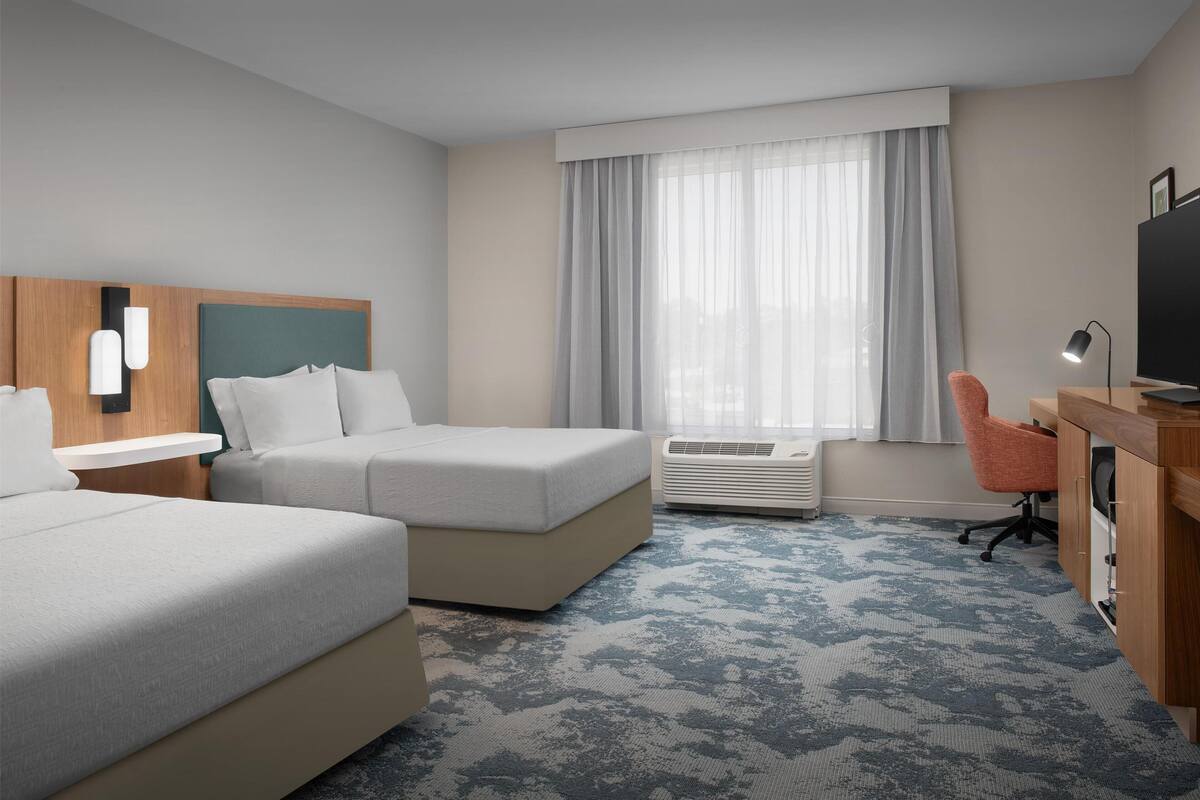 Une chambre d’hôtel avec deux lits, un bureau avec une chaise, une télévision et une grande fenêtre avec des rideaux.