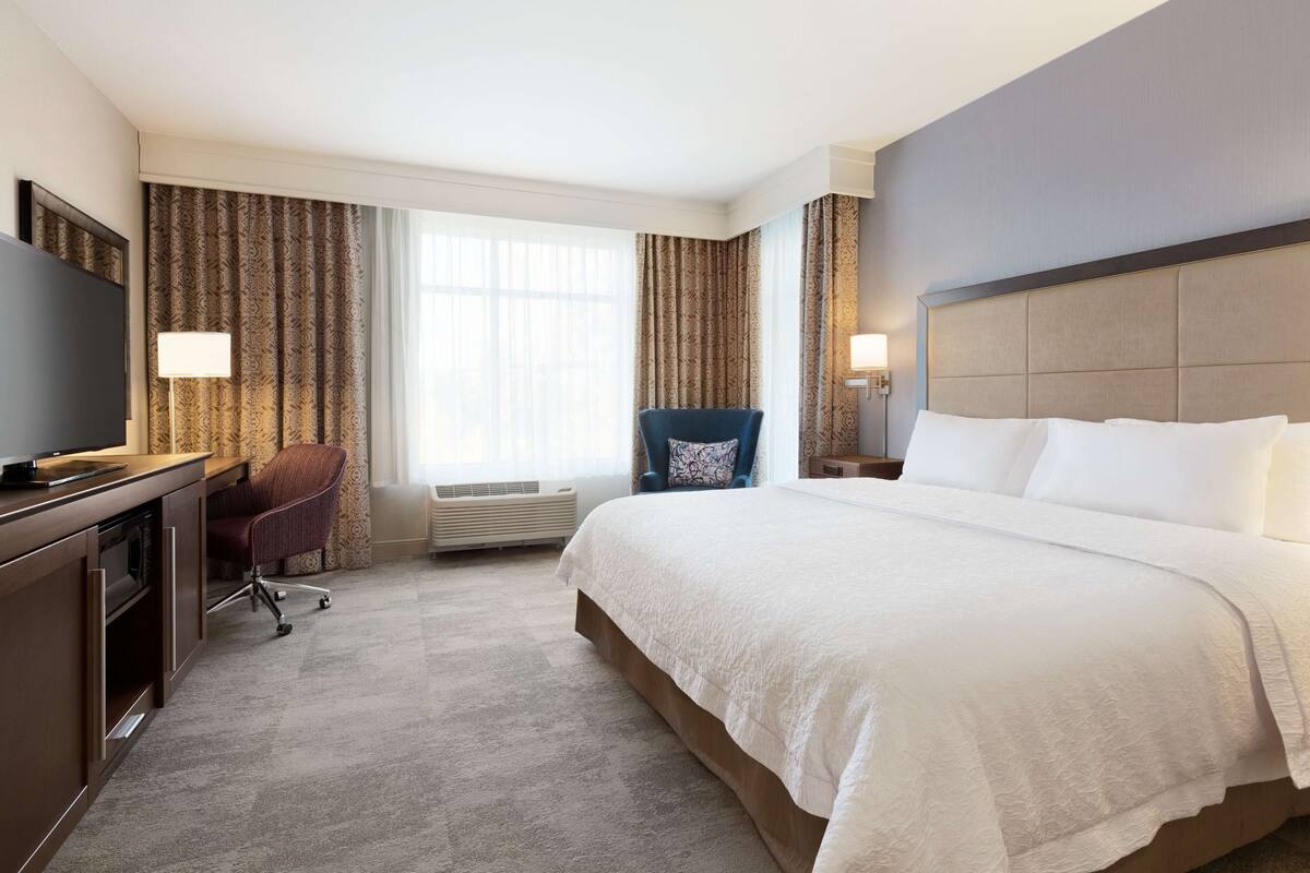 A hotel room with a large bed, a desk with a flat-screen TV, a chair, and a window with curtains.