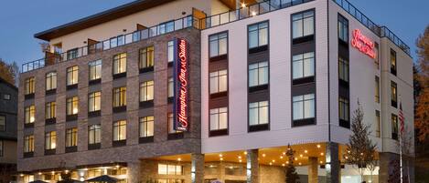 A multi-story hotel with a prominent 'Hampton Inn & Suites' sign, large windows, and a welcoming entrance with outdoor seating.