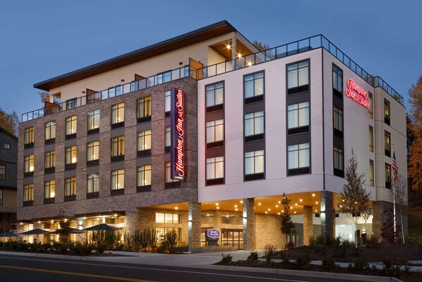 A multi-story hotel with a prominent 'Hampton Inn & Suites' sign, large windows, and a welcoming entrance with outdoor seating.