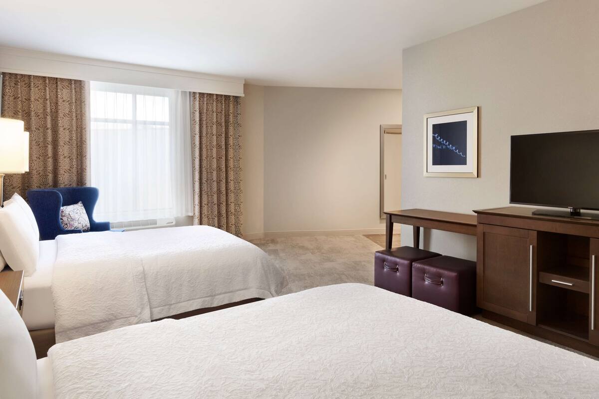 A hotel room with two beds, a desk, a television, and a window with curtains.