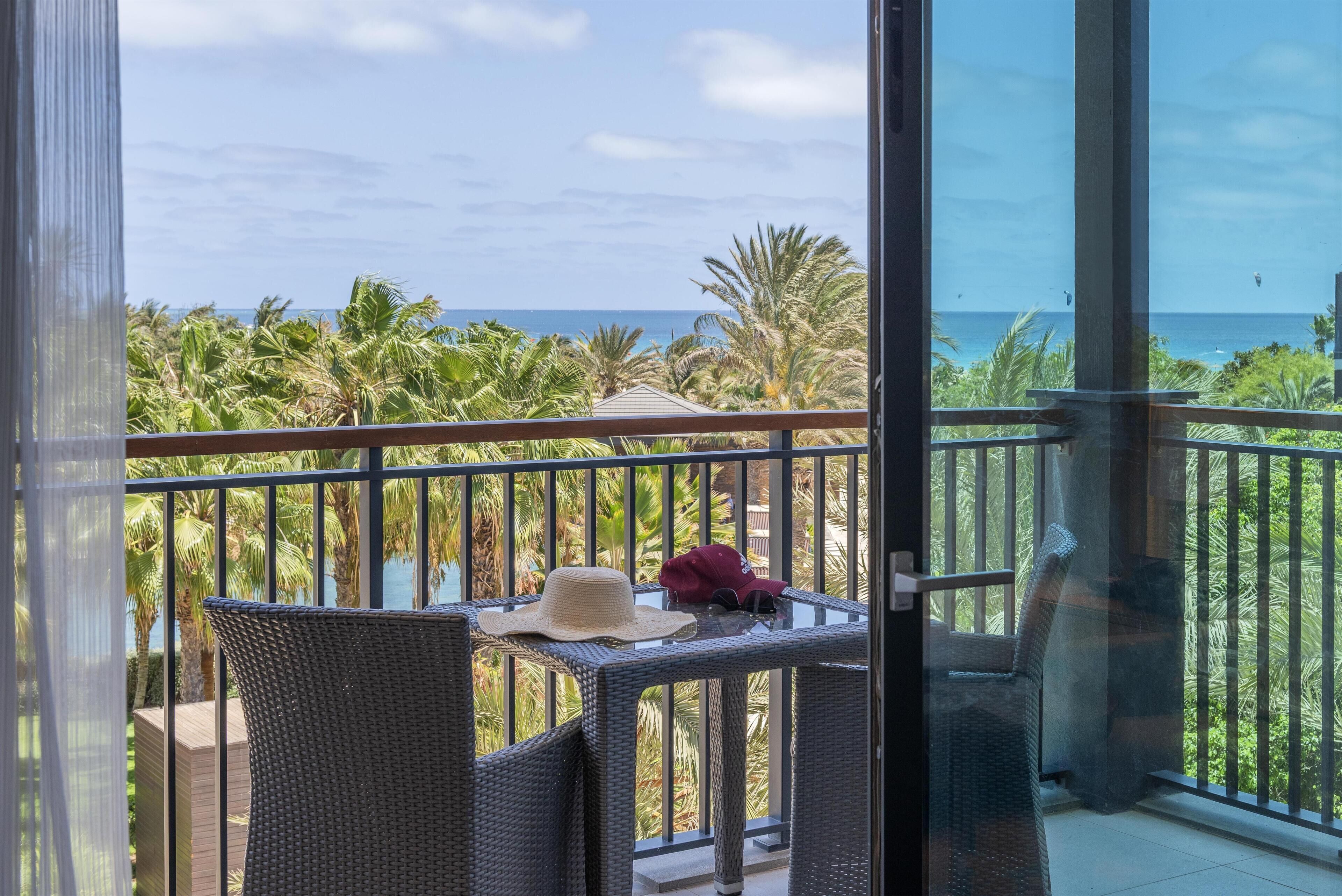 A balcony with wicker furniture, a hat, and a cap, overlooking a palm tree-lined view.