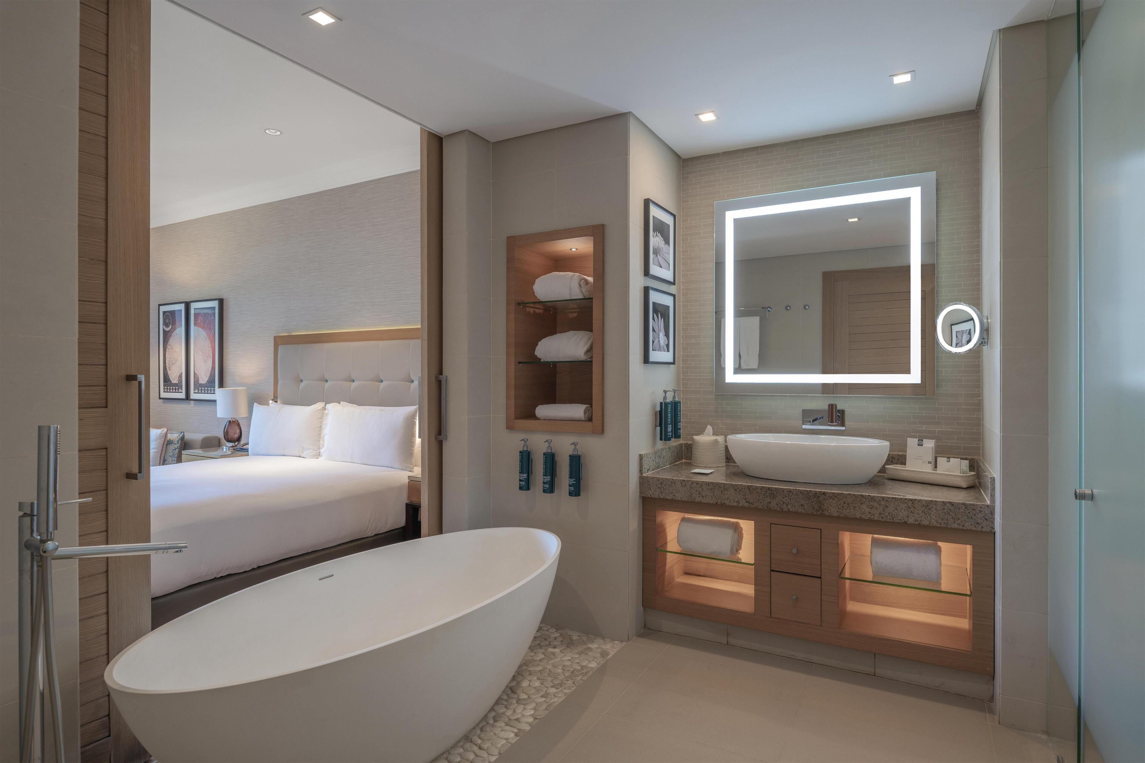 A modern bathroom with a large bathtub, a separate shower area, a vanity with a mirror, and a bed in the adjacent room.