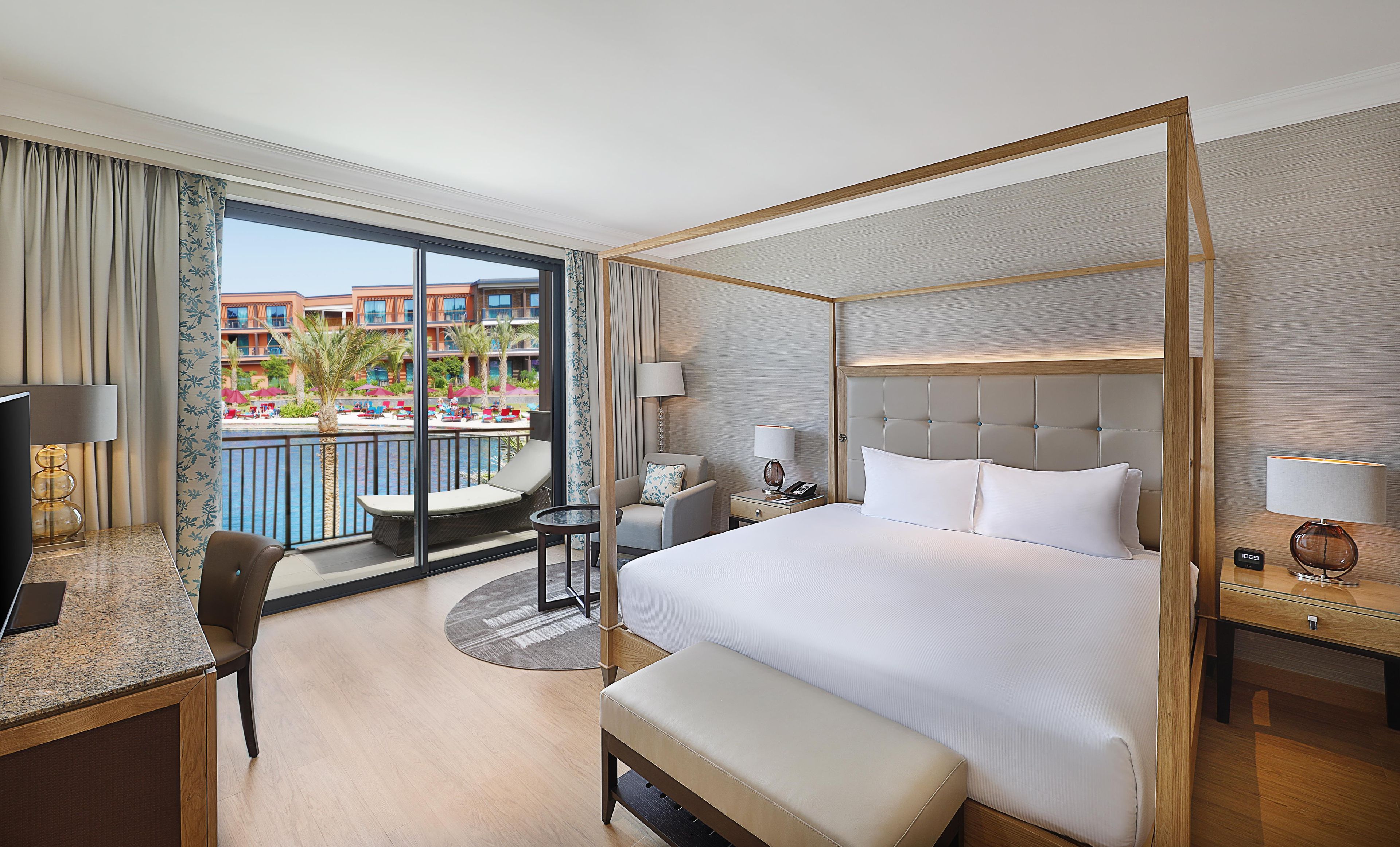 A hotel room with a four-poster bed, a desk with a chair, a small table, and a view of a pool and buildings.