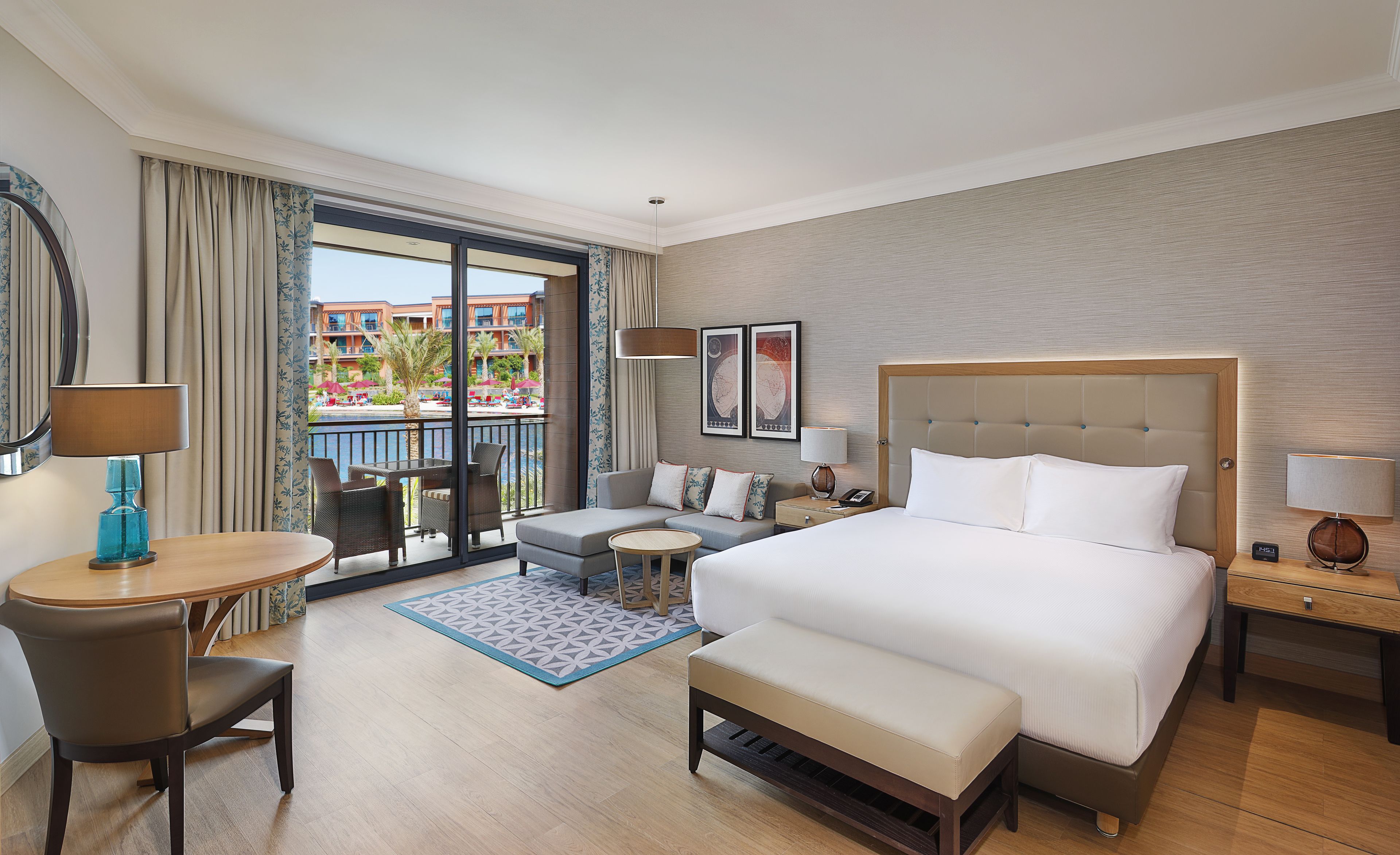 A hotel room with a large bed, a desk, a chair, a sofa, and a balcony with a pool view.